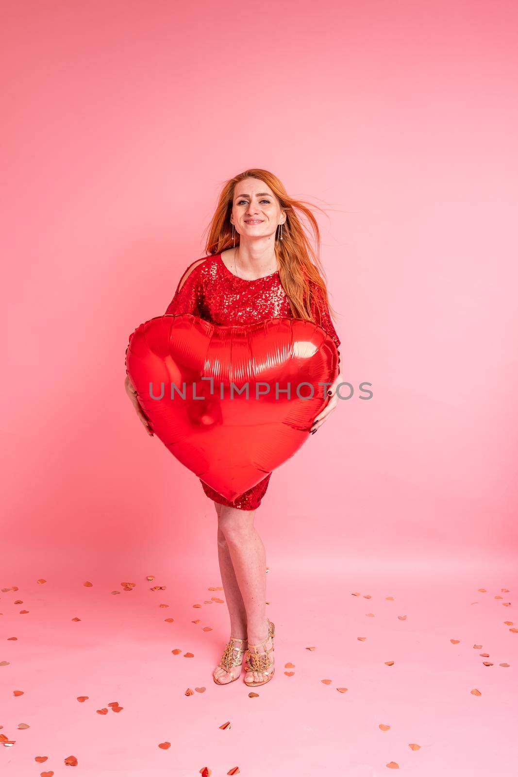 Beautiful redhead girl with red heart baloon posing. Happy Valentine's Day concept. Studio photo of beautiful ginger girl dancing on pink background.