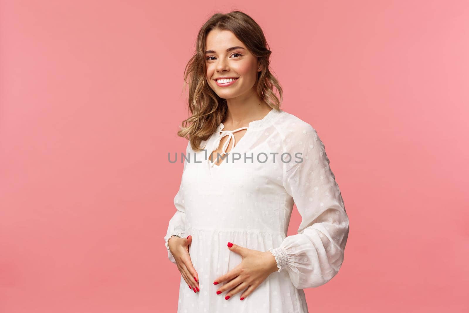 Maternity, women and beauty concept. Tender, cute smiling blond woman in white dress feeling happiness and love, touching belly as expecting child, being pregnant, pink background by Benzoix