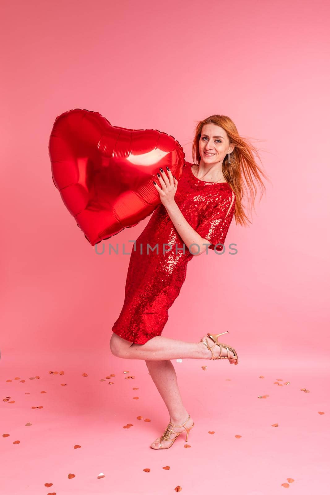 Beautiful redhead girl with red heart baloon posing. Happy Valentine's Day concept. Studio photo of beautiful ginger girl dancing on pink background.