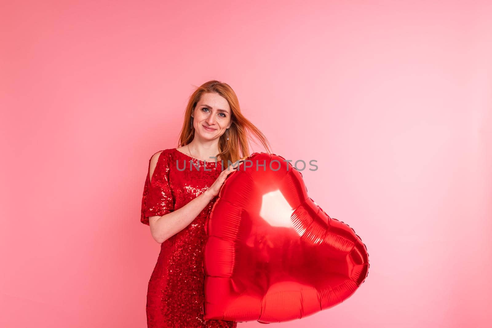 Beautiful redhead girl with red heart baloon posing. Happy Valentine's Day concept. Studio photo of beautiful ginger girl dancing on pink background.