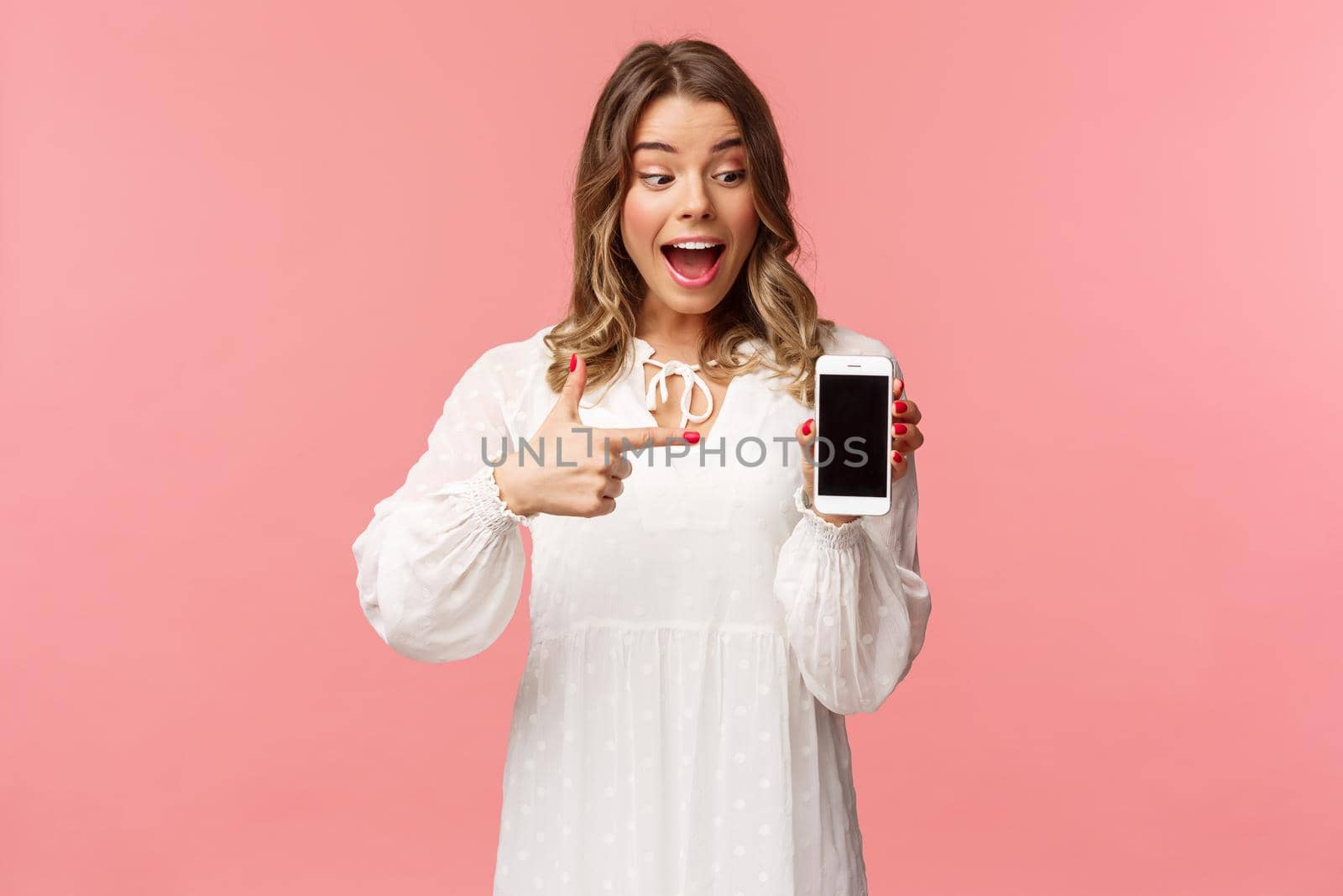 Portrait of excited and amazed, happy blond female in white dress, pointing finger at mobile phone screen, looking at smartphone with pleased and surprised smile, stand pink background.