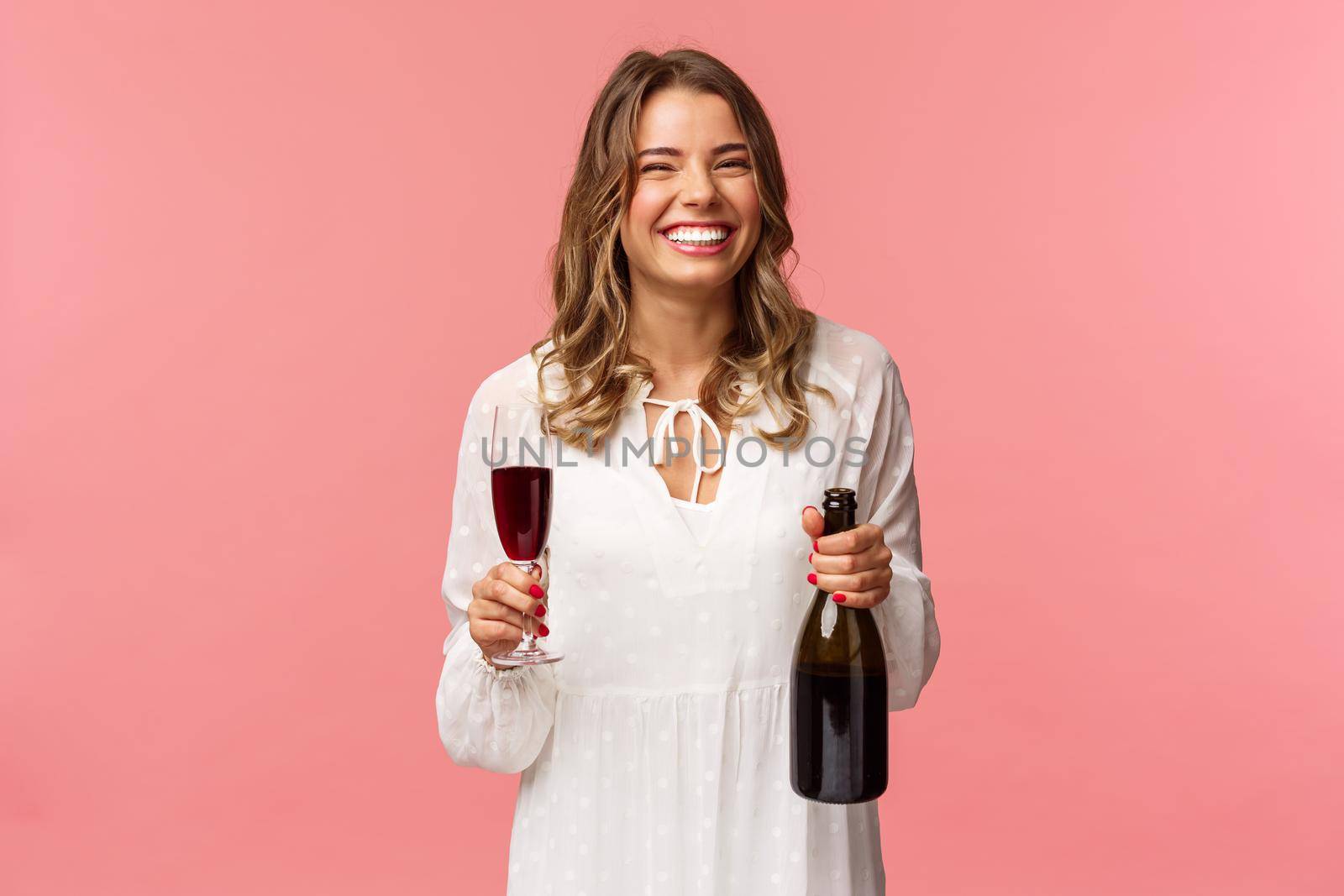 Holidays, spring and party concept. Portrait of happy and carefree european blond female celebrating in white dress, holding bottle champagne or wine, drinking from glass and laughing.