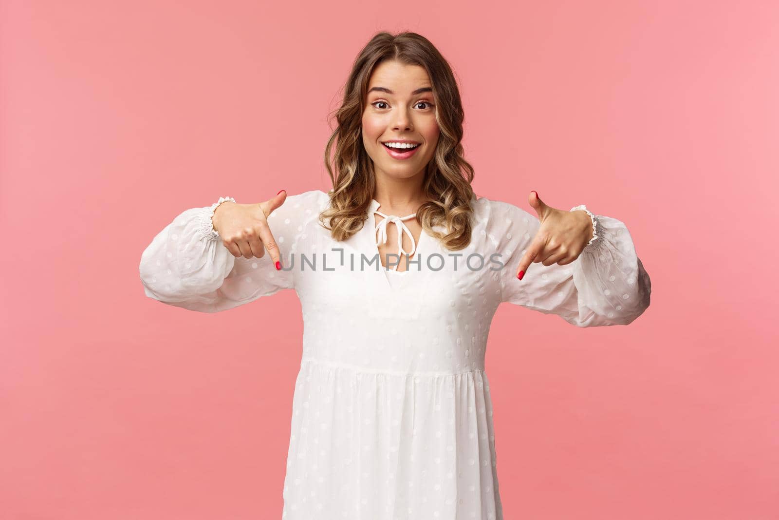 Portrait of enthusiastic upbeat young blond woman pointing fingers down to invite you check-out product, showing bottom advertisement, smiling camera cheerful, spring concept, pink background.