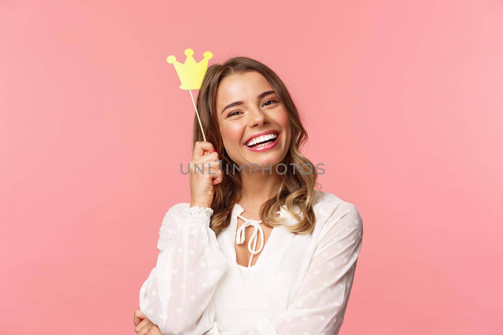 Spring, happiness and celebration concept. Close-up portrait of charming smiling, lovely blond girl holding small queen crown on stick, laughing joyfully, feel empowered and happy, pink background by Benzoix