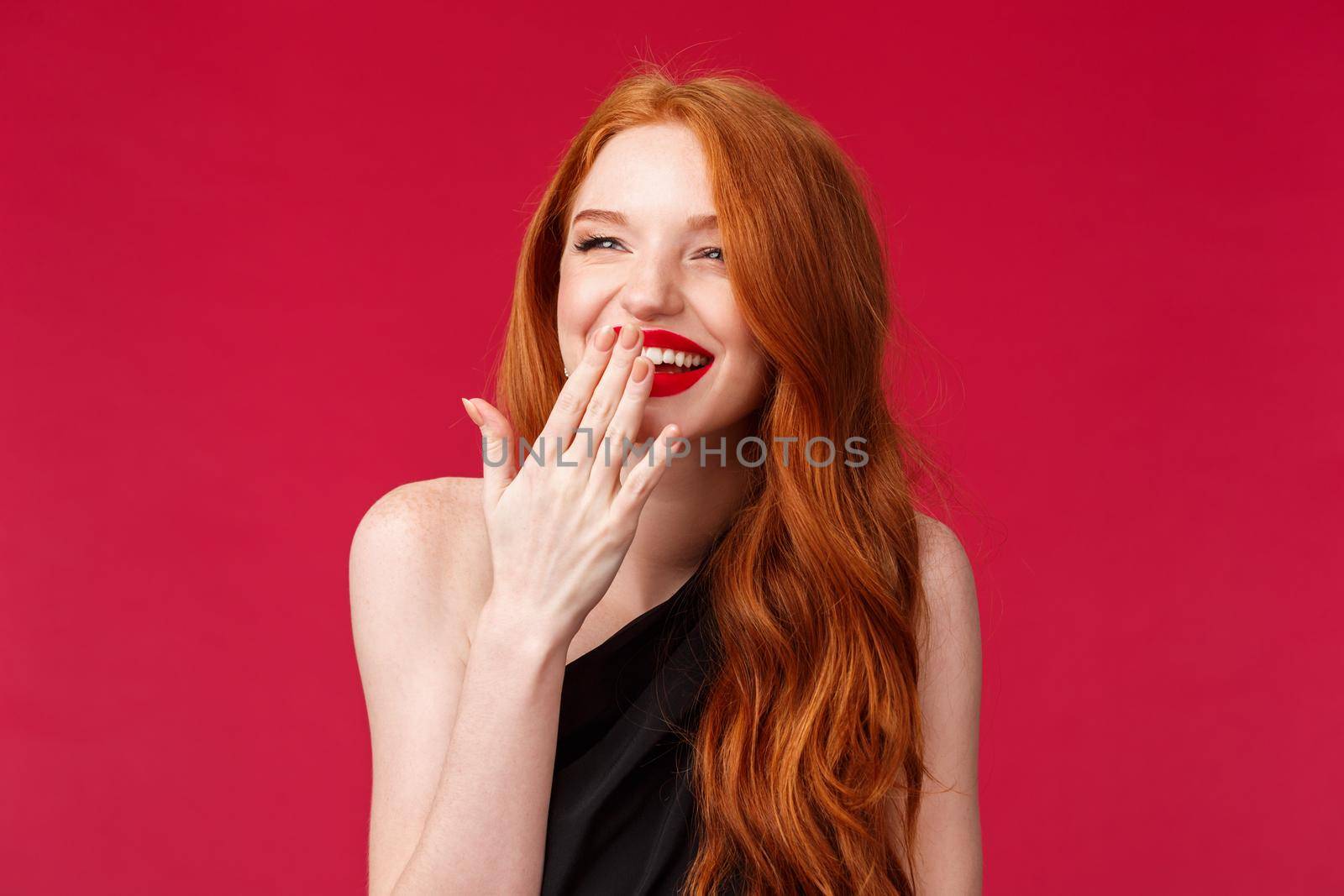 Close-up portrait of pretty elegant young european woman with long curly red hair, black dress and evening makeup, laughing over something funny looking away and cover smile with palm.