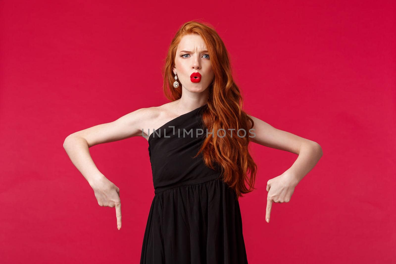 Celebration, events, fashion concept. Portrait of frustrated and upset serious redhead elegant woman in trendy black dress, complaining grimacing dissatisfied, pointing fingers down.