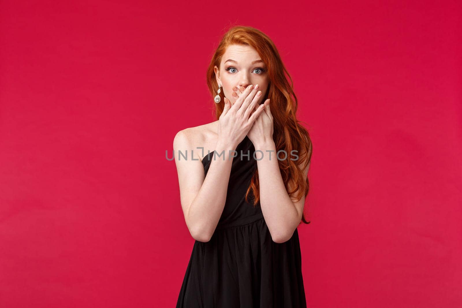 Fashion, luxury and beauty concept. Portrait of shocked, speechless redhead female in black evening dress, gasping and cover mouth impressed, stare camera startled, red background by Benzoix