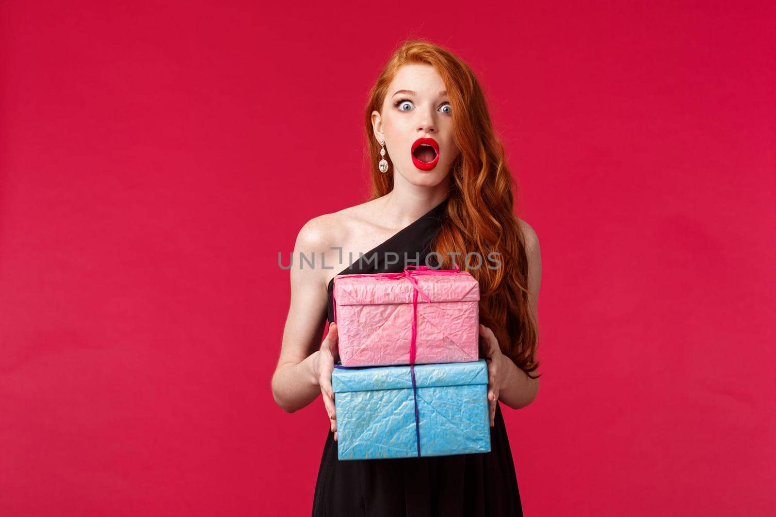 Celebration, holidays and women concept. Portrait of amazed and excited, ambushed young redhead female in black dress, drop jaw holding two cool gifts, stand red background.