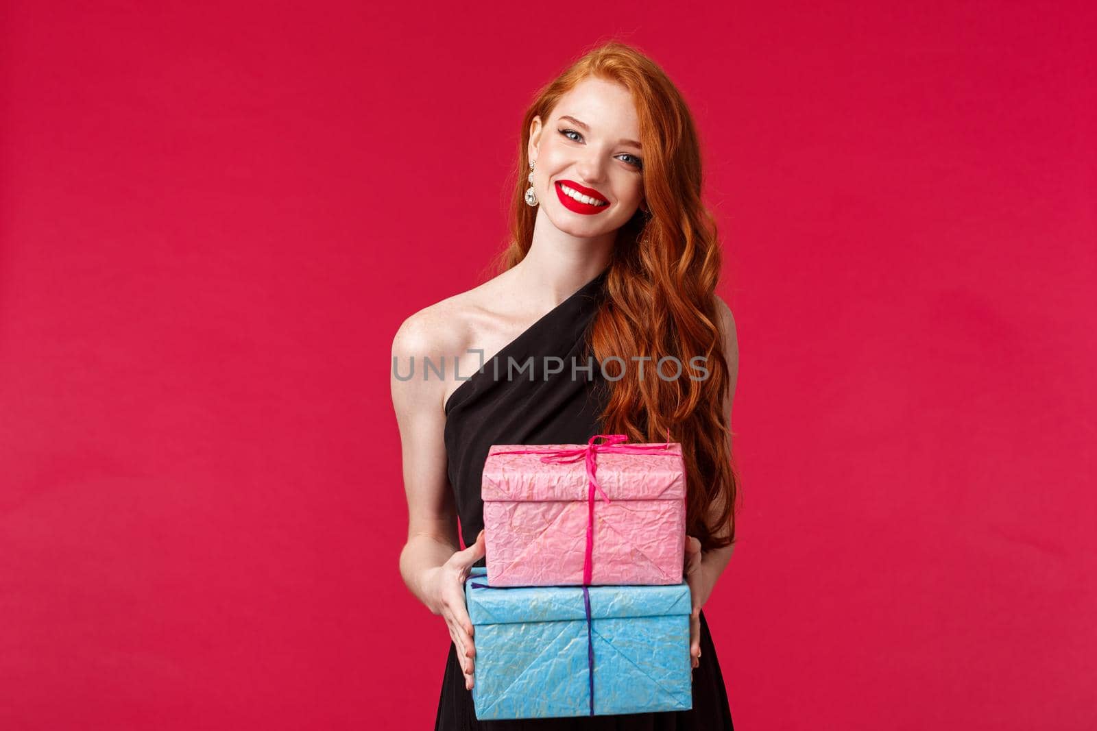 Celebration, holidays and women concept. Charming redhead female in black evening dress, red lipstick, give presents to birthday girl, party owner, holding two gifts and smiling cheerful.