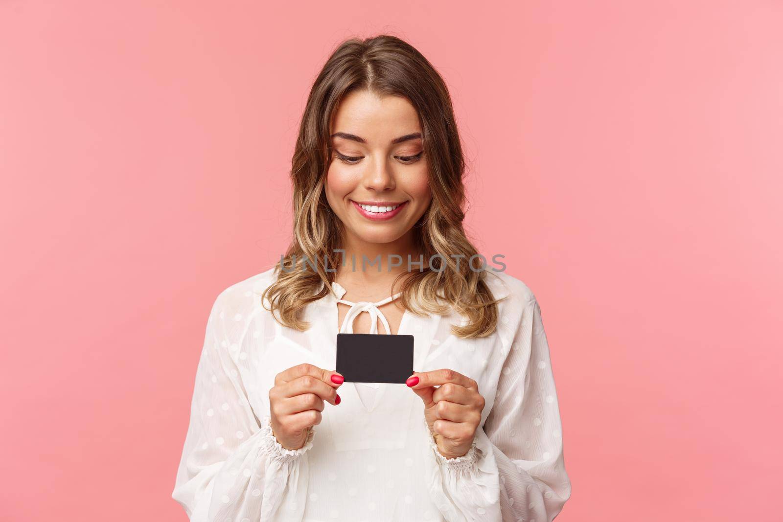 Close-up portrait of excited and amused blond girl in white dress, holding credit card and smiling thrilled, cant resist temptation to buy something, waste money online shopping, pink background by Benzoix