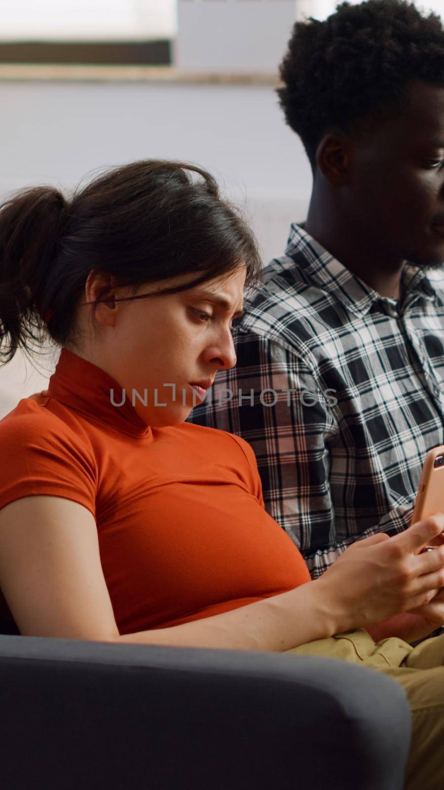 Modern interracial people holding smartphones on couch while sitting together in living room. Young mixed race couple using devices and technology for entertainment as lifestyle