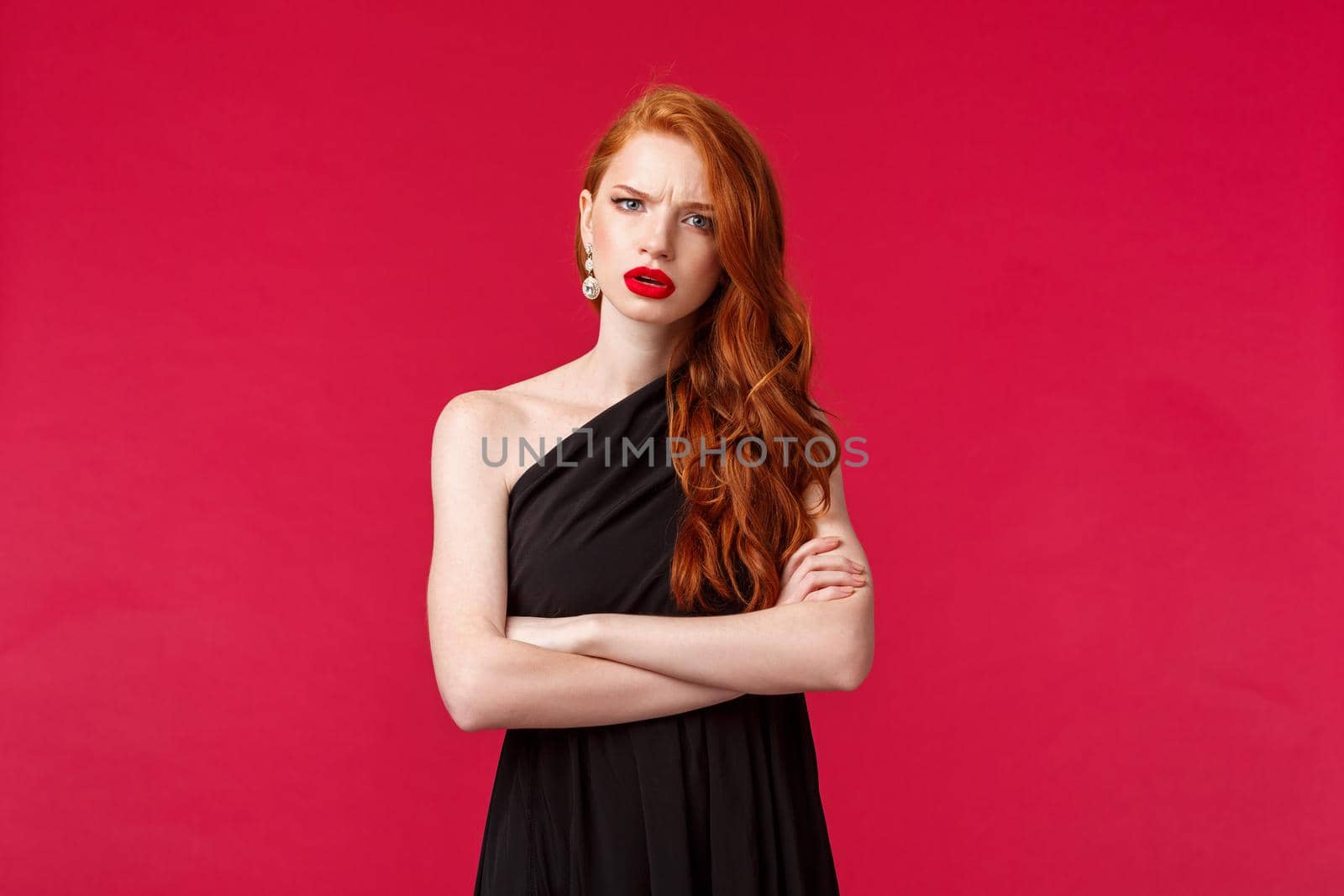 Portrait of serious and concerned, displeased young redhead woman in black dress, cross hands chest, frowning at camera wtih accusation and judgement, listening to something frustrating.