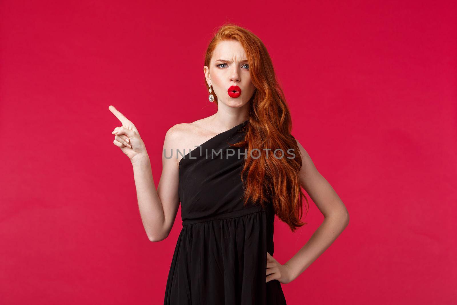 Beauty, fashion and women concept. Disappointed and dubious young redhead woman pointing finger left and frowning at camera upset, complain or being frustrated, wear black evening dress by Benzoix