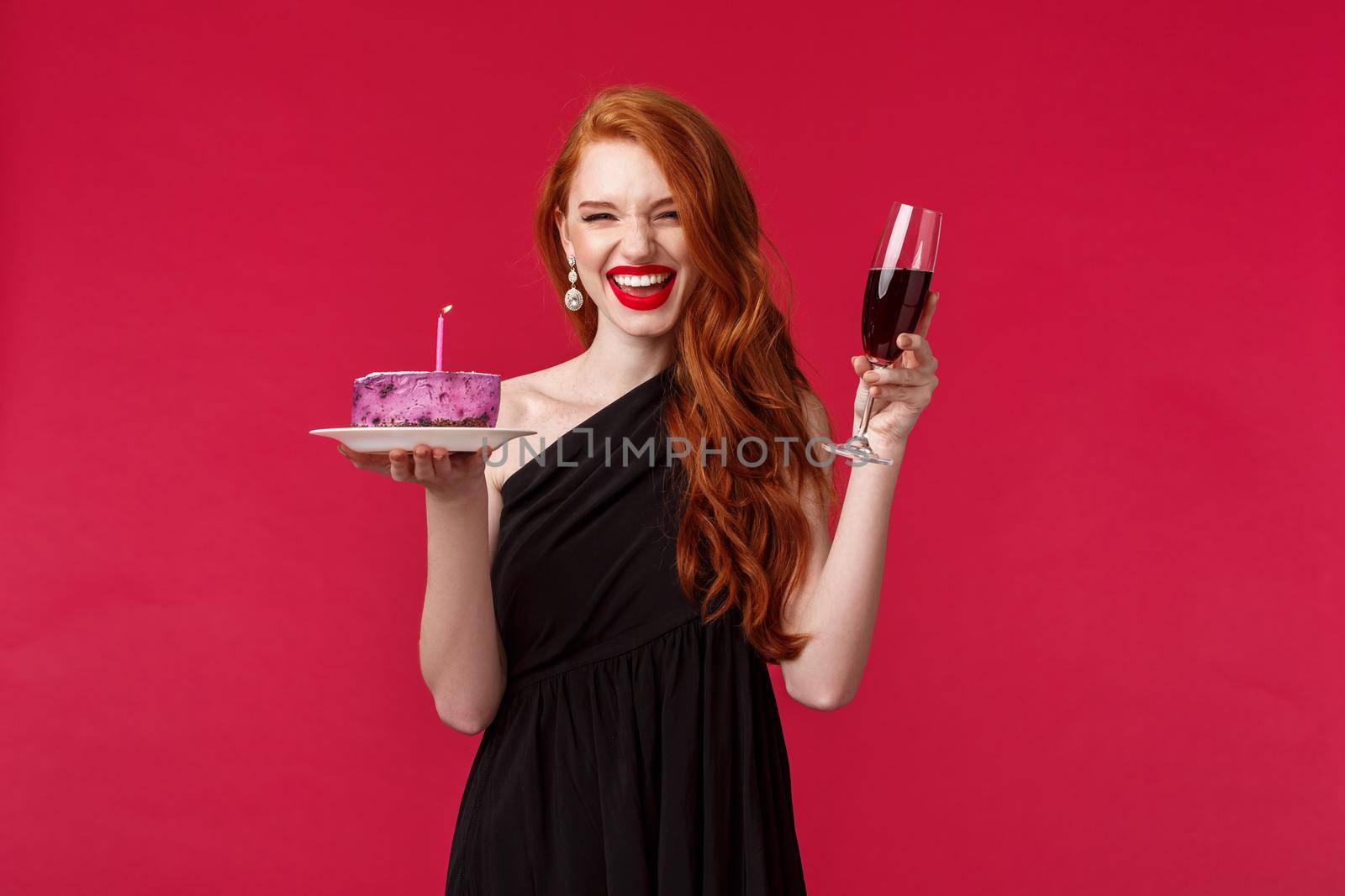 Portrait of excited laughing gorgeous redhead woman having fun at b-day party, holding glass of wine and birthday cake with lit candle, making wish, celebrating over red background.