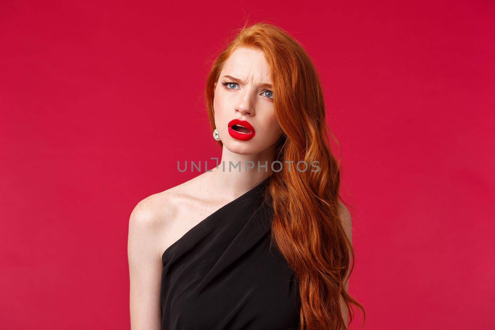 Close-up portrait of perplexed and confused young redhead woman seeing something strange, squinting and frowning as looking left troubled identify what is this, stand red background.