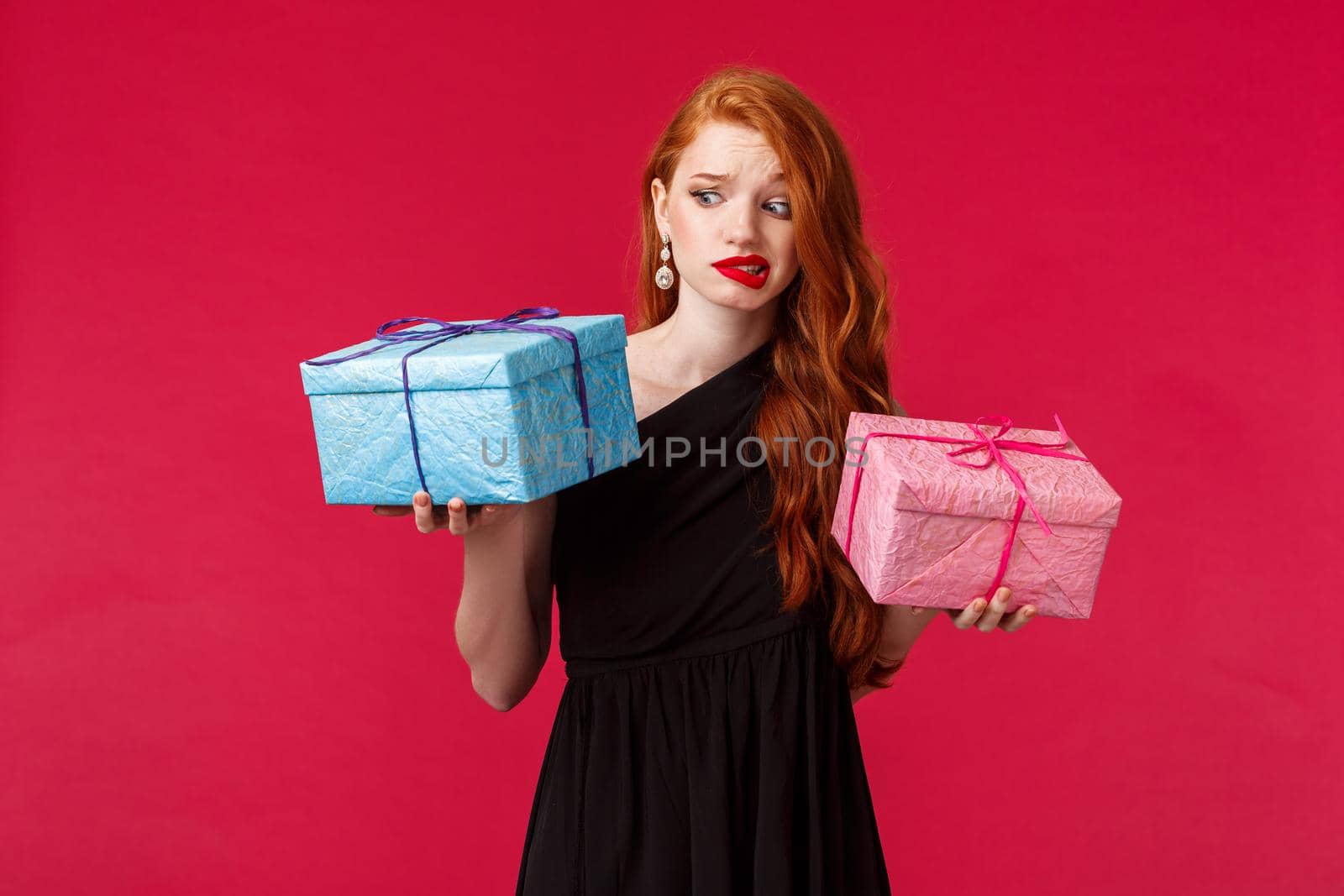 Celebration, holidays and women concept. Portrait of skeptical and unsure attractive caucasian redhead woman receive strange gift, grimacing at something strange or ugly, red background.