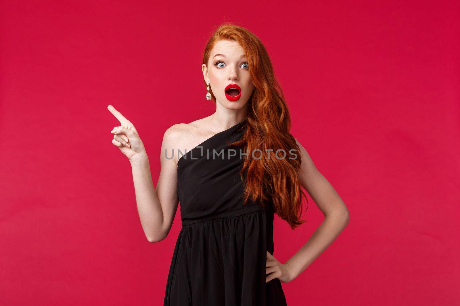 Beauty, fashion and women concept. Elegant young surprised woman in red lipstick, wear stylish black dress, gasping amazed at camera as pointing finger left want to know what happened.