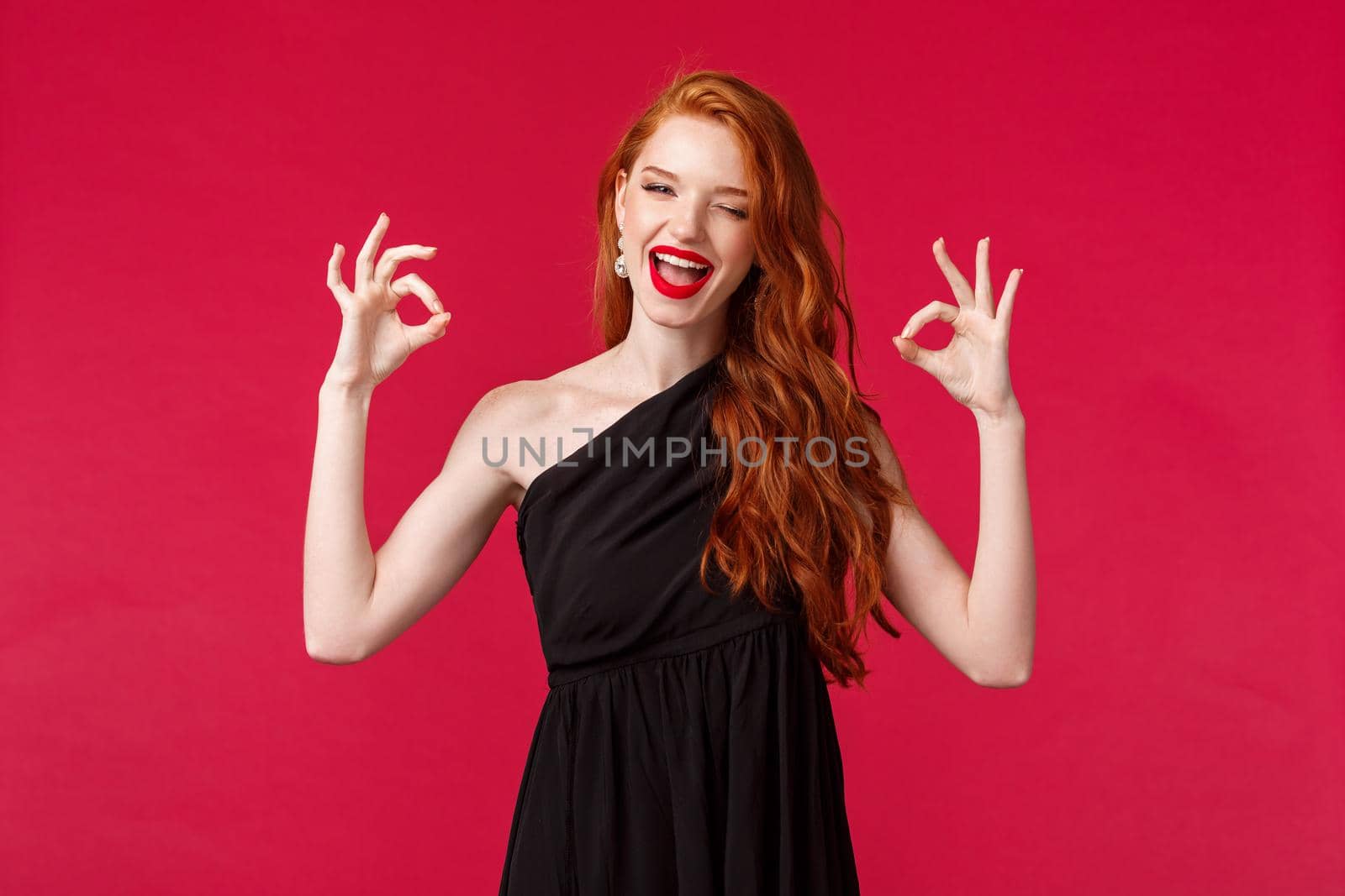 Fashion, luxury and beauty concept. Portrait of cheerful sassy caucasian female with ginger hair, black dress and makeup, show okay signs, no problem, agree or approve something, red background.