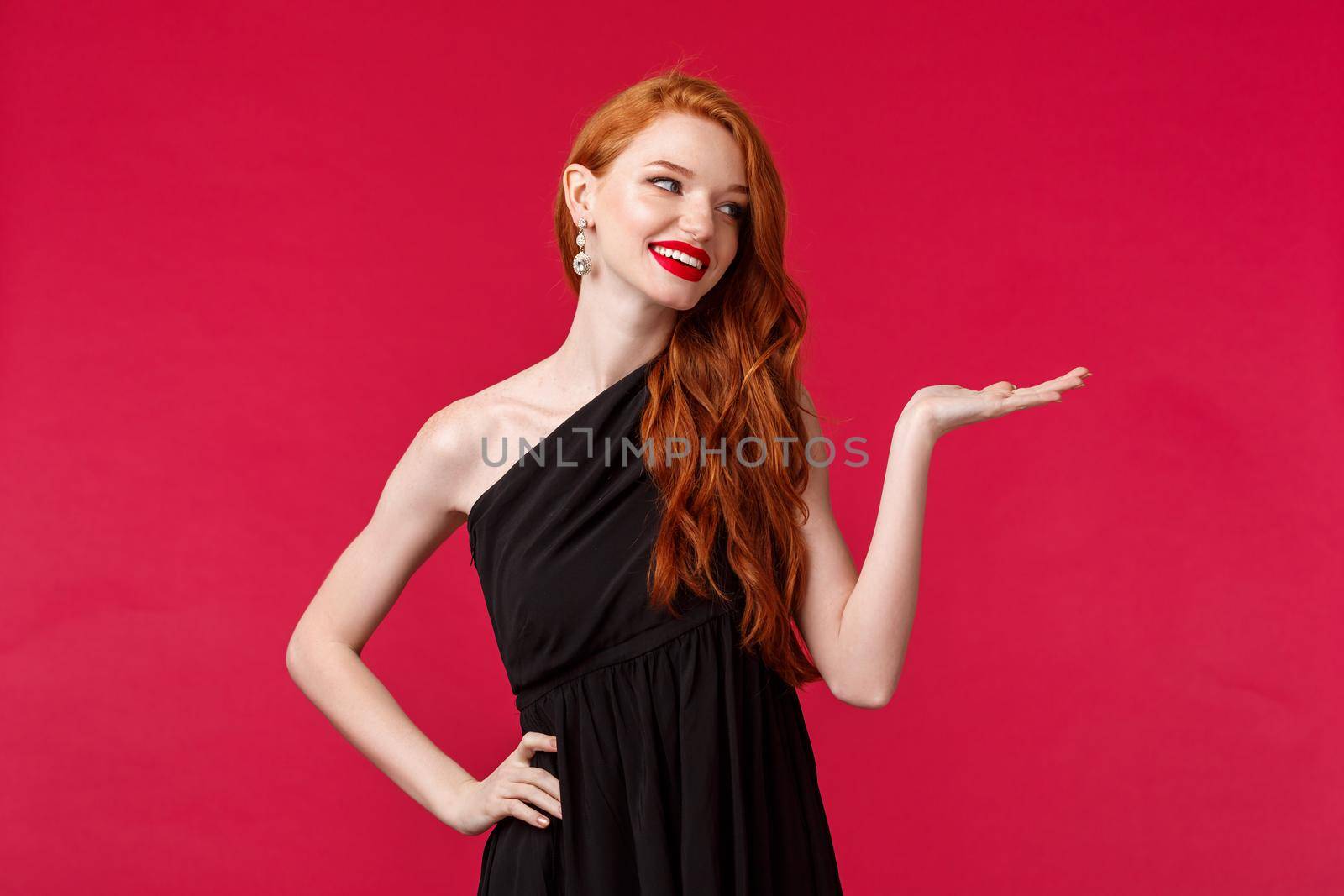 Portrait of luxurious good-looking redhead woman in black evening dress, attend event, look at blank space smiling, holding product on arm, promotion of company, red background.