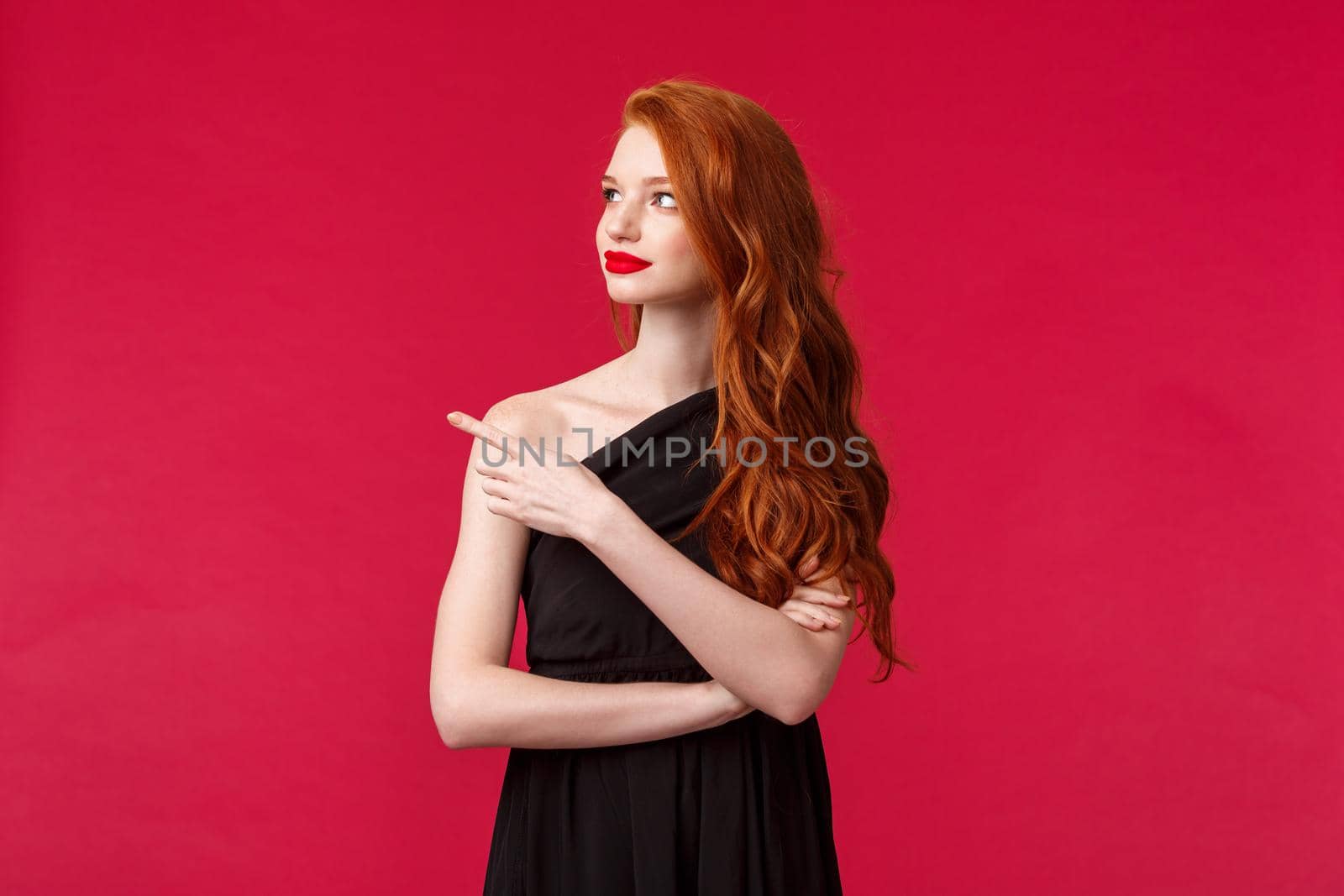 Celebration, events, fashion concept. Portrait of elegant young redhead slim woman in black stylish dress, wear makeup red lipstick, look and pointing left with curious interested face by Benzoix