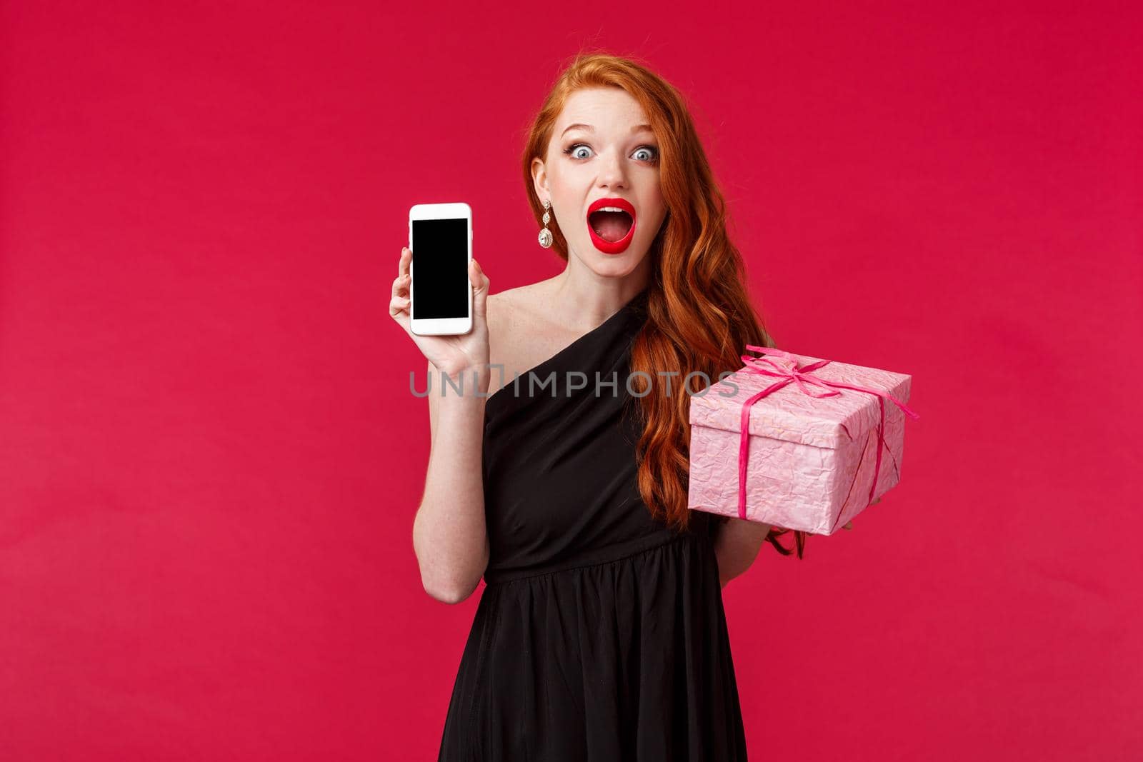 Portrait of amazed and excited redhead woman holding mobile phone, showing smartphone screen, application or online store, hold gift box, receive awesome present, gasping stunned camera.