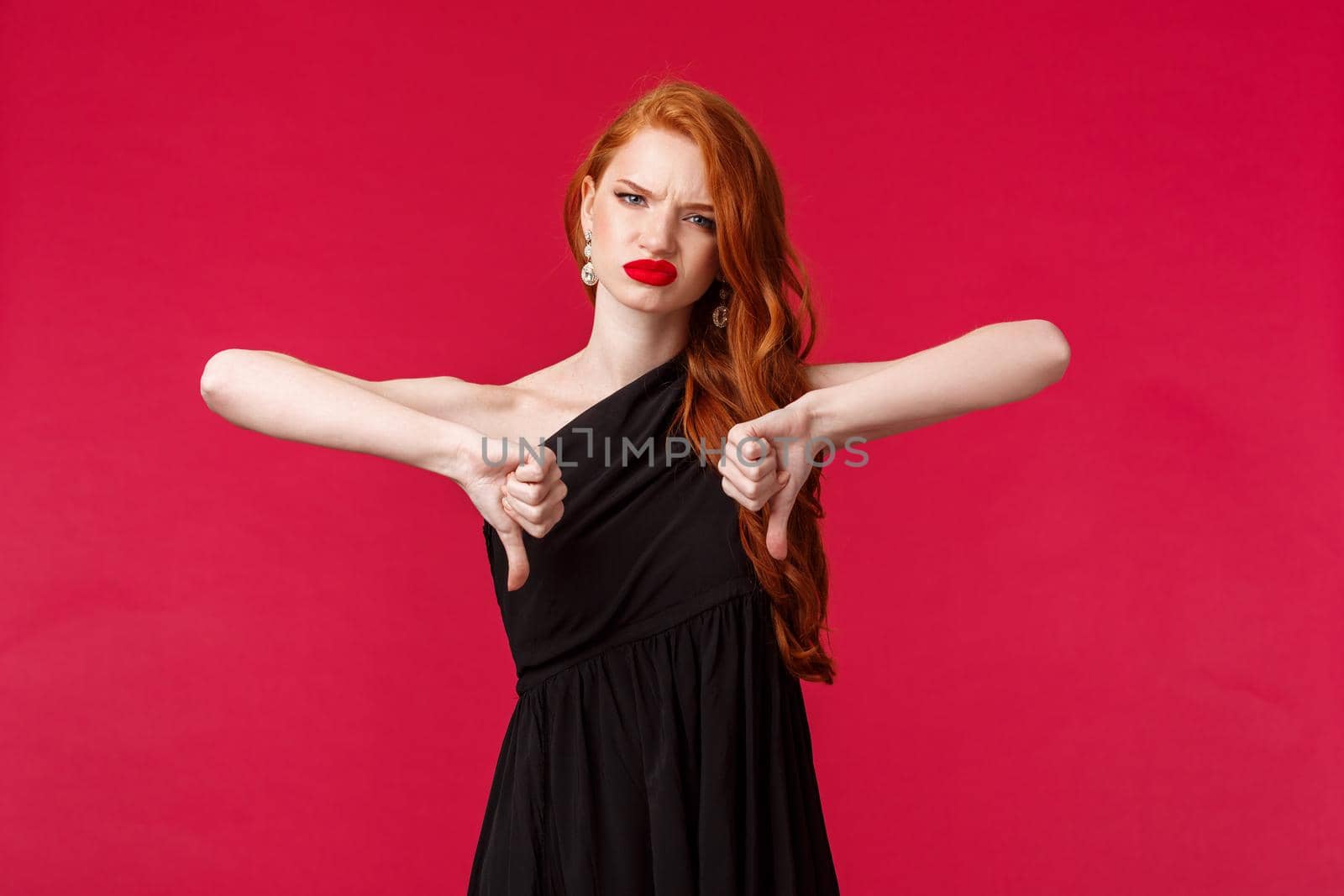 Fashion, luxury and beauty concept. Portrait of disappointed, unsatisfied gorgeous young redhead woman in black dress disagree with bad person, show thumbs-down grimacing in disapproval.