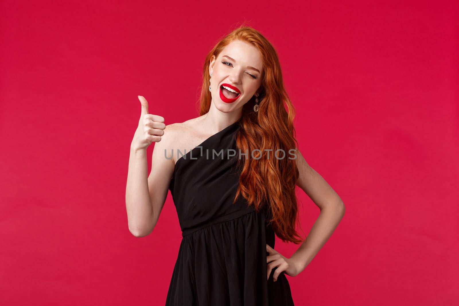 Fashion, luxury and beauty concept. Portrait of sassy good-looking elegant woman with red hair, black dress, wink at camera seductive, show thumb-up in like, approval gesture, red background.
