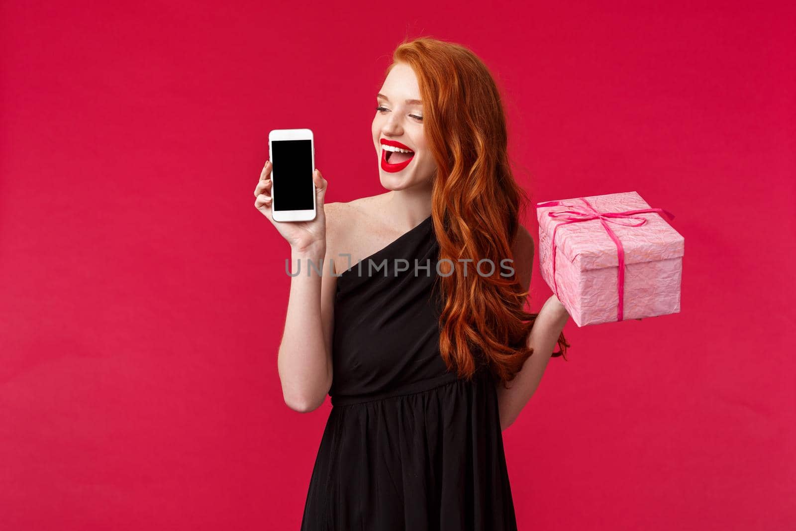 Portrait of excited beautiful woman with ginger hair, wear black stylish dress, holding at looking at mobile phone display with happy pleased expression, hold birthday gift, present pink box.