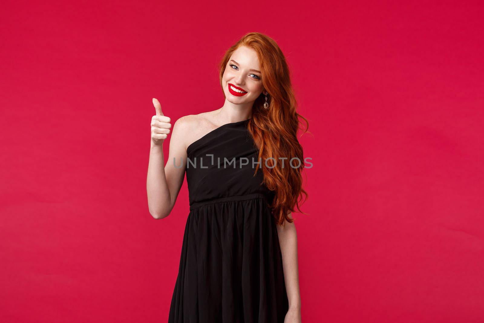 Elegance, fashion and woman concept. Portrait of satisfied charming young elegant redhead woman in black stylish dress over red background, show thumbs-up in approval or like sign.