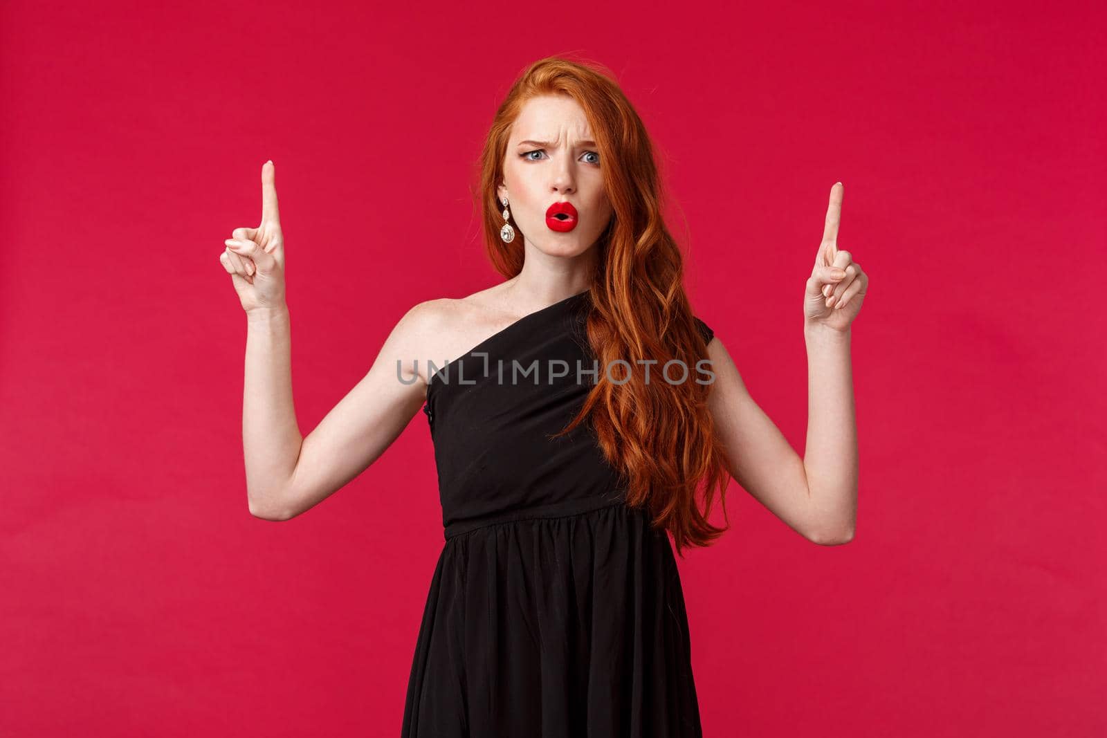 Celebration, events, fashion concept. Portrait of frustrated and puzzled redhead woman in red lipstick, black elegant dress, frowning disappointed, pointing up at something bad.