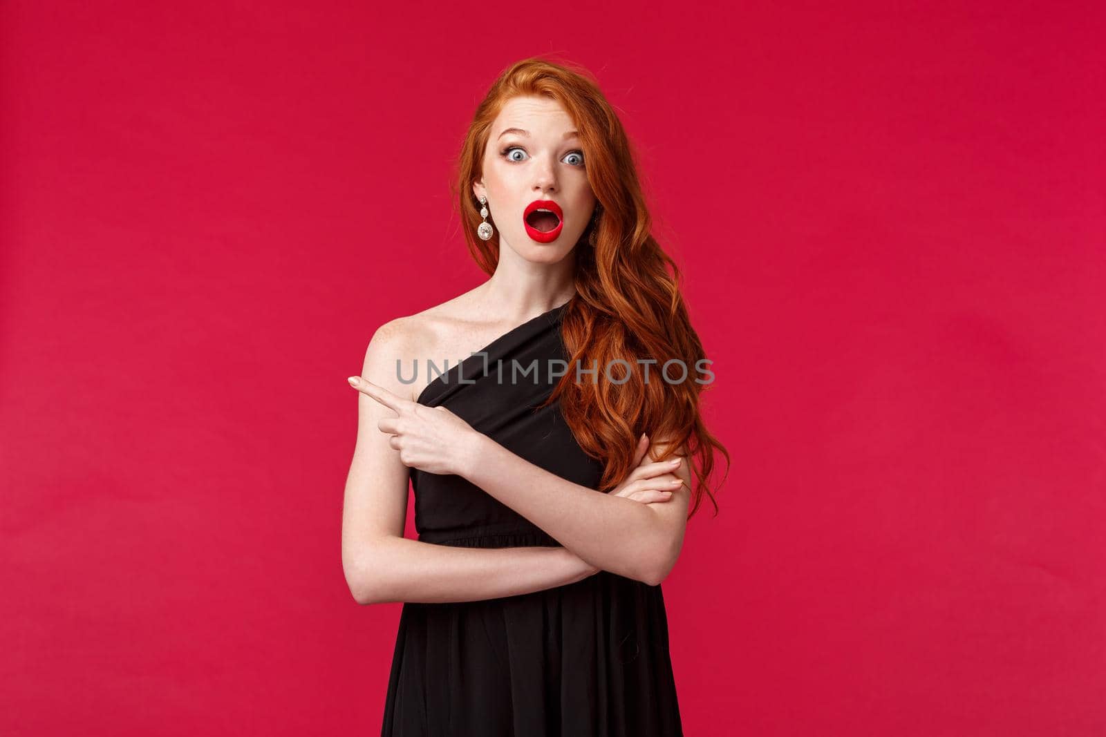 Elegance, fashion and woman concept. Portrait of ambushed and surprised young redhead woman, girl in black dress looking shocked, gossiping pointing finger left, red background by Benzoix