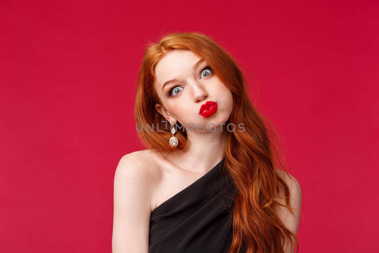 Close-up portrait of silly and funny pretty redhead woman in red lipstick, makeup and black evening dress, tilt head pouting like wish and fold lips mwah, stand red background.