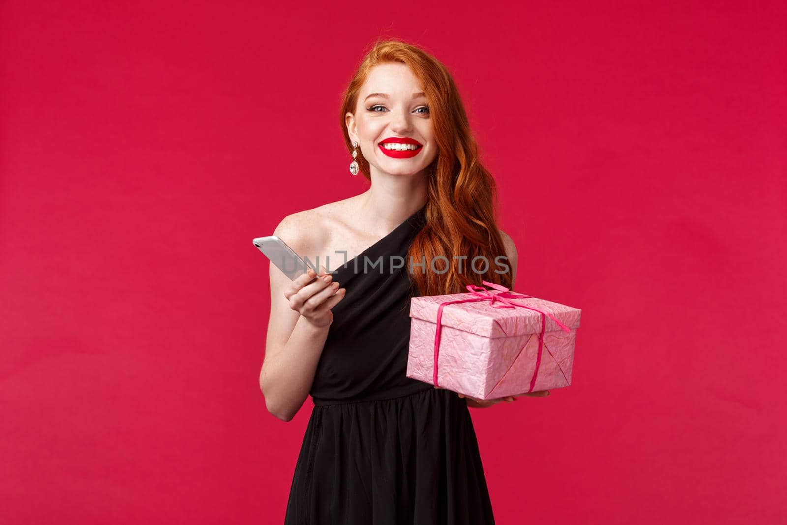 Portrait of happy, excited attractive woman with ginger hair, celebrating birthday, holiday with presents, receive gift, hold box and smartphone laughing smiling camera, red background.