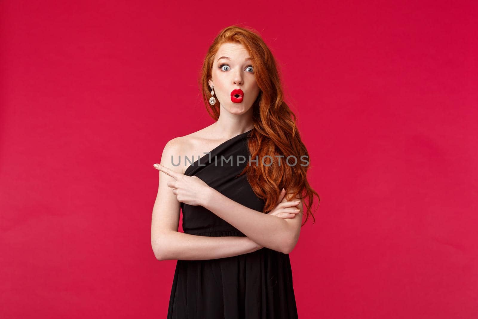 Elegance, fashion and woman concept. Portrait of amused and excited redhead attractive woman in black elegant dress, red lipstick and evening makeup, asking question curious pointing finger left.