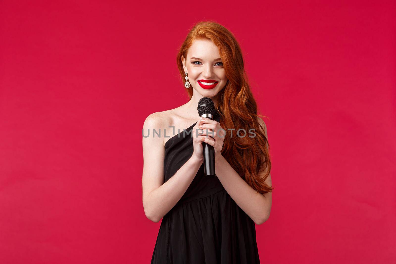 Portrait of elegant beautiful young woman with red long hair wearing evening dress, holding microphone, performer singing for clients in restaurant, smiling sensually, red background.