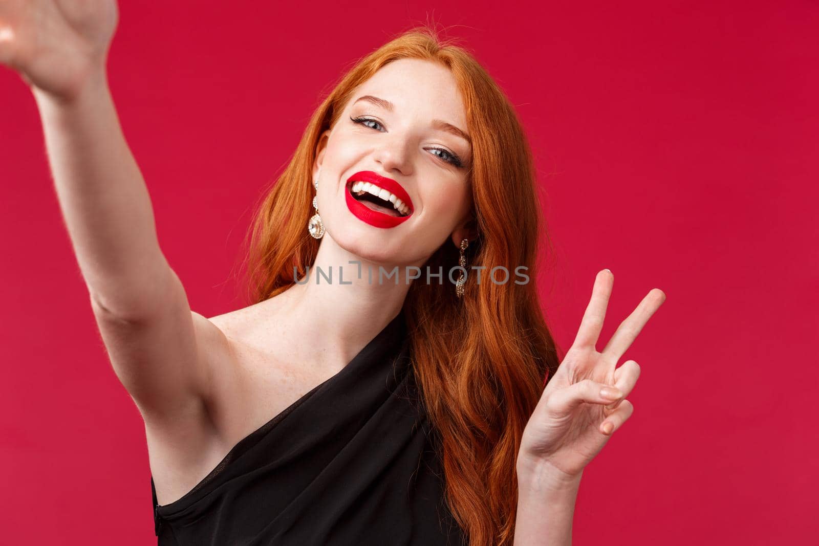 Close-up portrait of happy charming redhead woman enjoying party, taking selfie with peace sign and beaming smile, wear black evening dress and red lipstick, stand red background by Benzoix