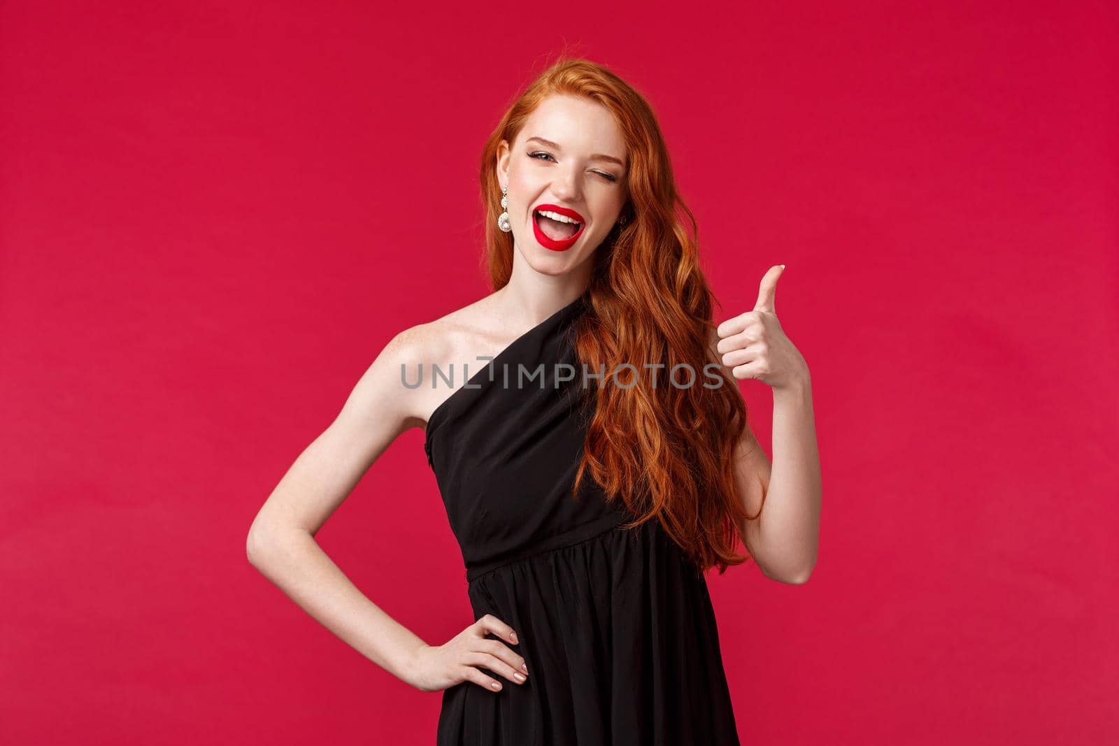 Fashion, luxury and beauty concept. Portrait of sassy gorgeous young redhead woman in black dress, wink at camera with thumb-up, recommend or approve something really awesome, likes party.