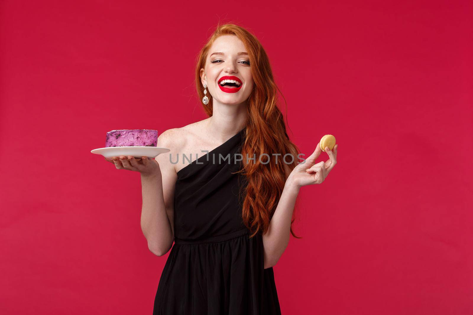 Portrait of beautiful carefree redhead woman in black dress, laughing over funny joke at party, holding cake on plate and cookie, eating delicious desserts, enjoying perfect birthday celebration.