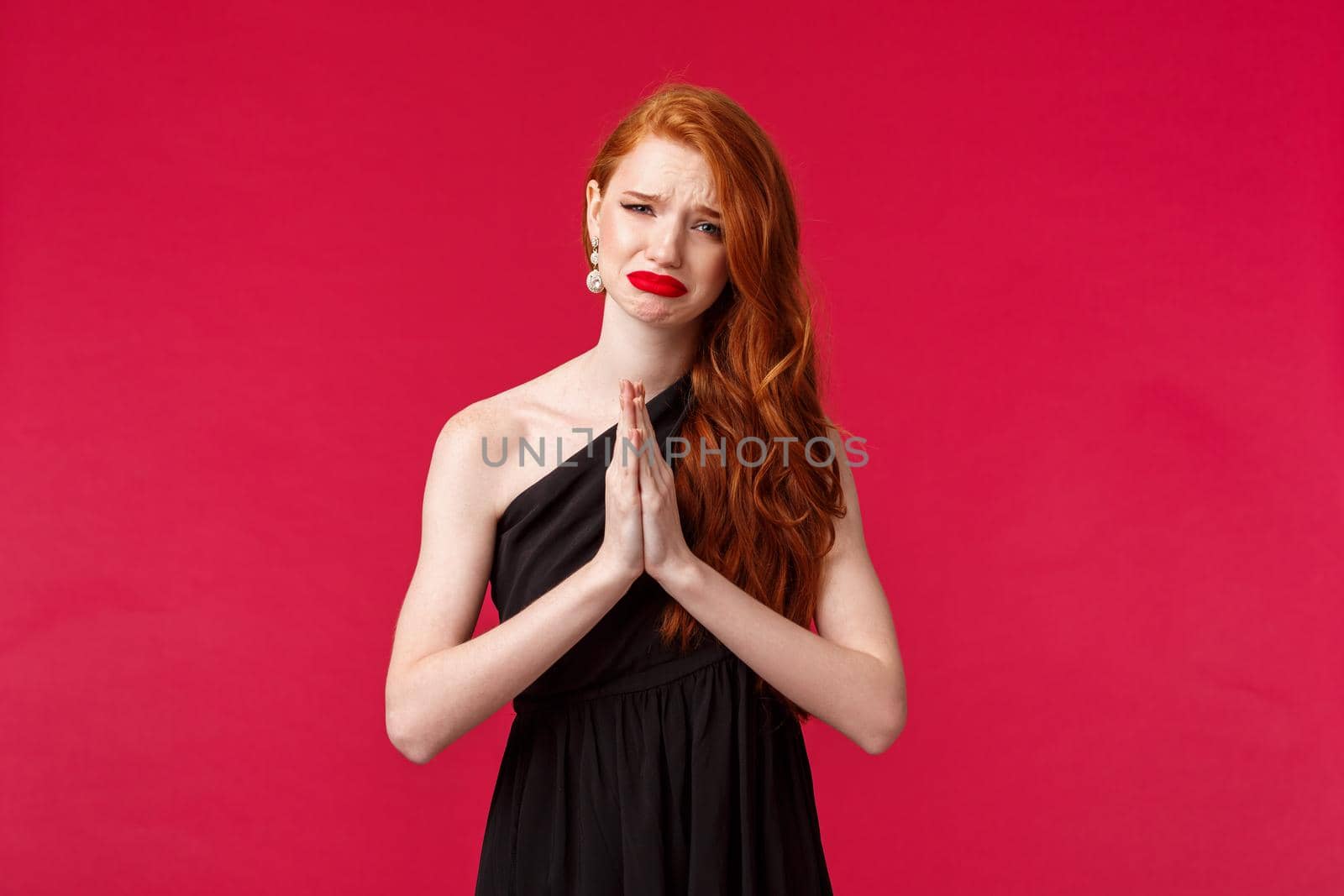 Portrait of upset and sad cute gloomy redhead woman in black evening dress, whining begging for help, need something badly, grimacing crying desperate, praying over red background.