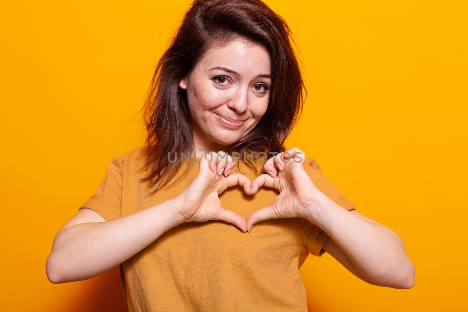 Positive adult making heart shape with fingers at camera. Portrait of romantic person doing love sign and symbol with hands, showing affection and emotion over orange background.