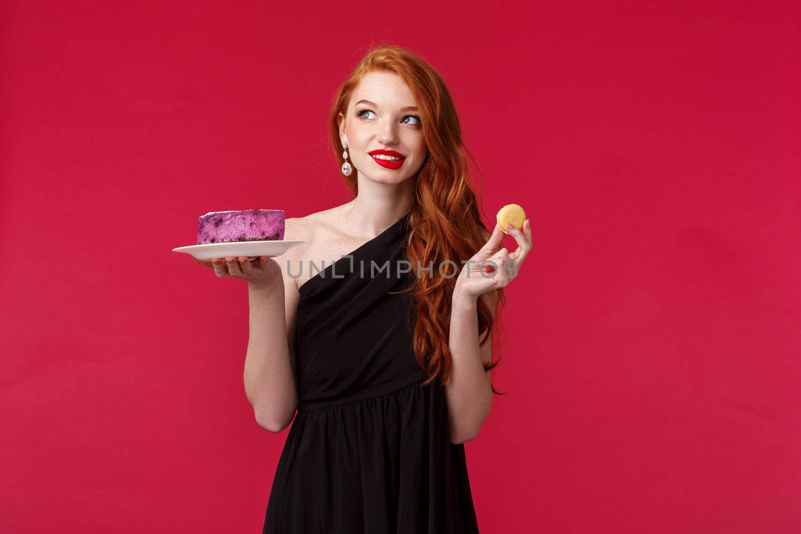 Thoughtful and dreamy gorgeous slim redhead woman in black dress thinking about eating dessert while taking care of her body, look away pondering and smiling, hold cookie and cake.