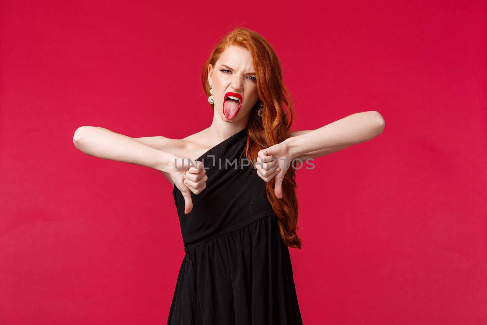 Fashion, luxury and beauty concept. Portrait of disappointed and bothered young redhead woman with long ginger hair, luxurious black dress over red background, show thumbs-down dislike.