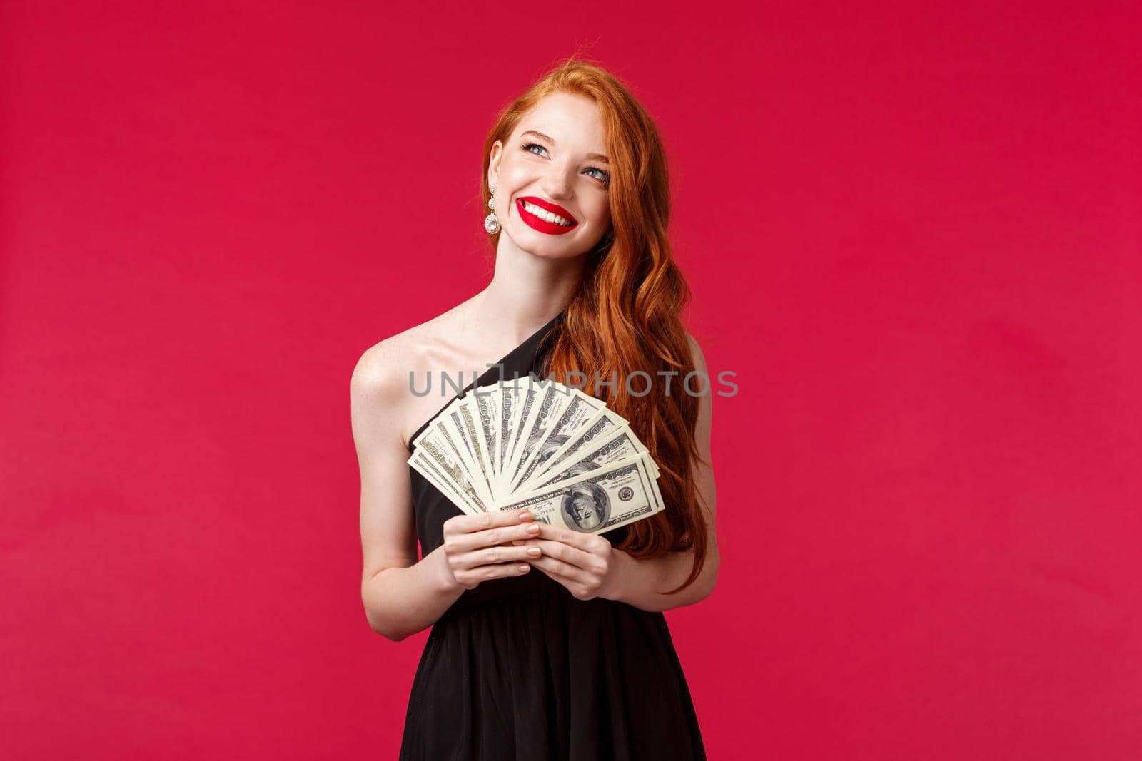 Luxury, beauty and money concept. Portrait of lucky and carefree rich attractive redhead woman in stylish evening dress, laughing happy looking up thinking, holding cash dollars, red background.