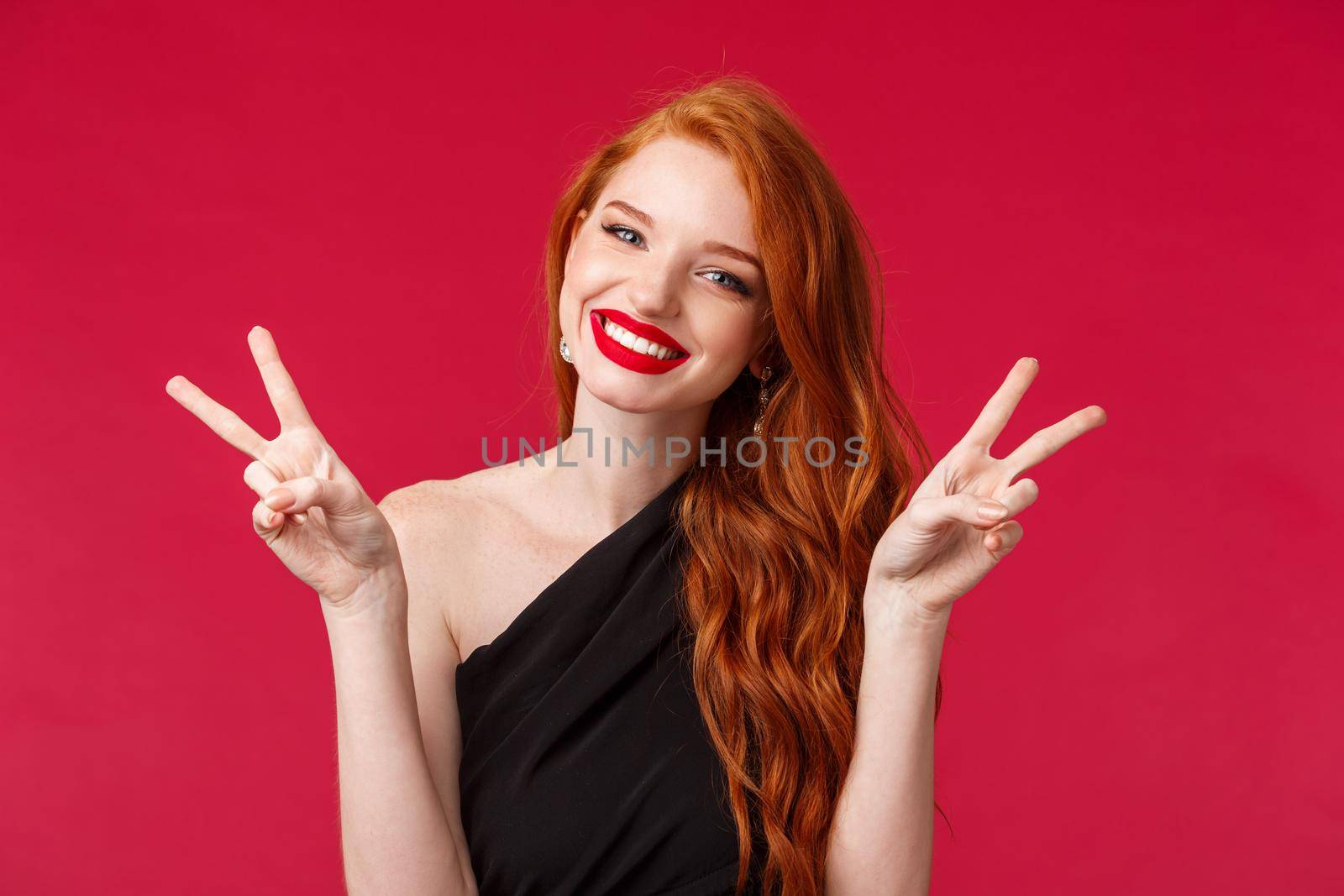 Close-up portrait of cheerful beautiful redhead woman in black dress, red lipstick, show kawaii peace signs, enjoying party, having fun, living a dream, standing red background upbeat.