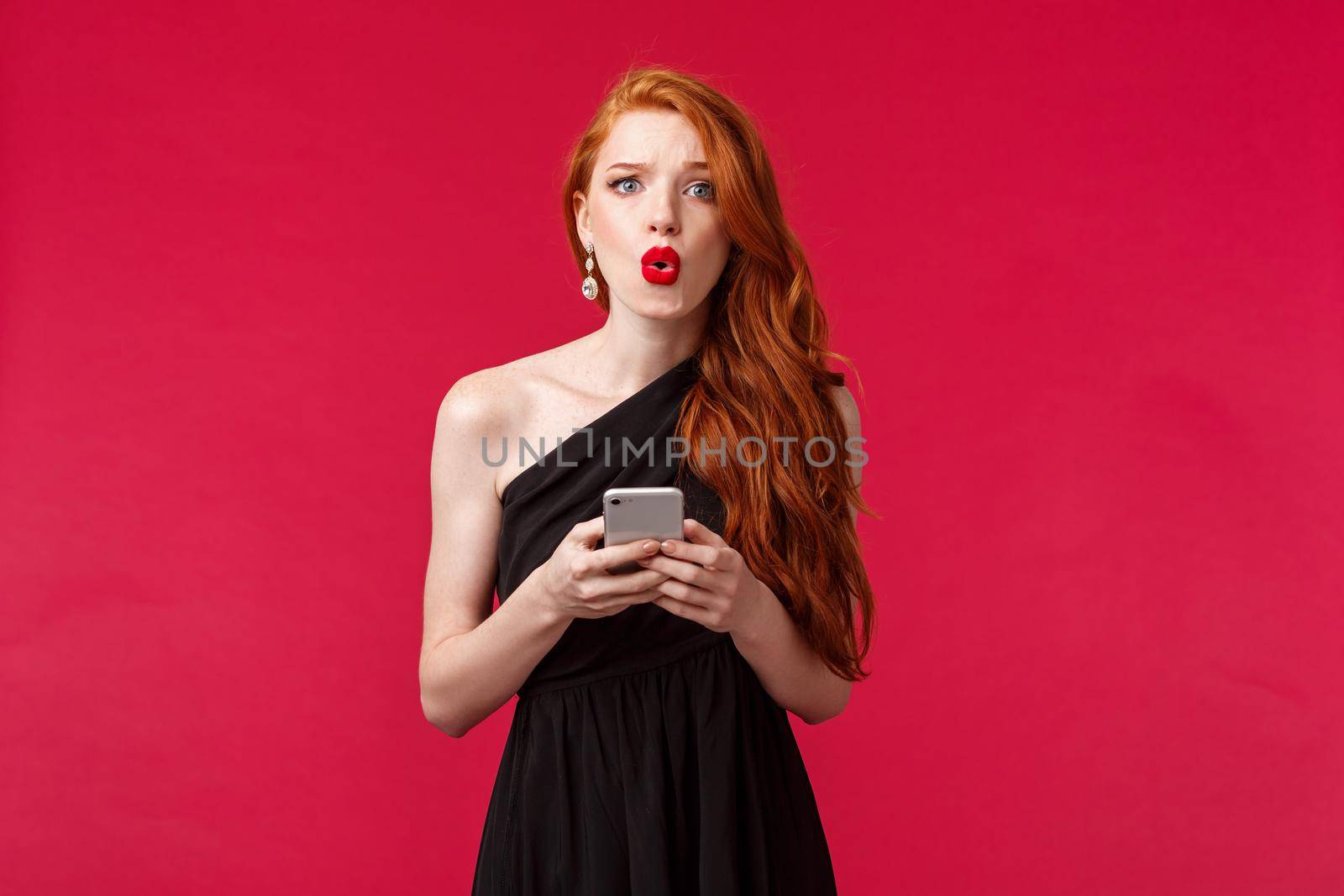 Portrait of concerned and confused young redhead woman being dumped by message on her prom night, look frustrated dont understand what happened, wear black dress, holding smartphone.