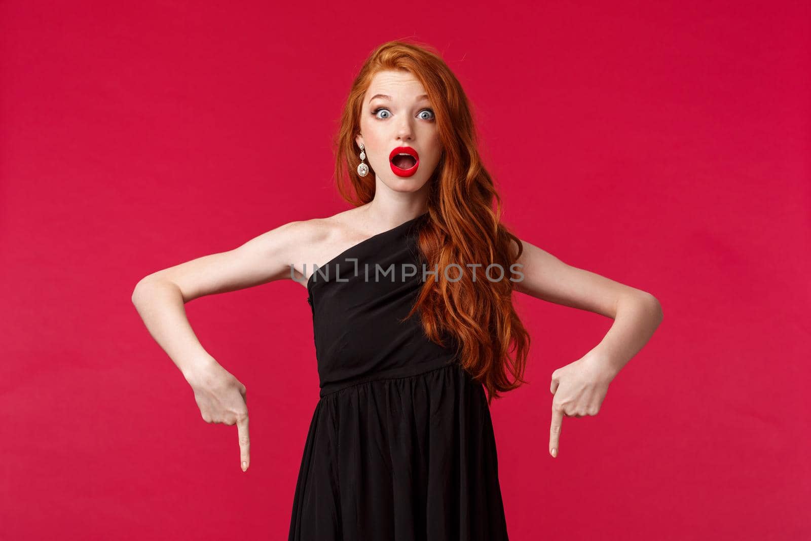 Celebration, events, fashion concept. Surprised and amazed redhead young woman in black dress attend awesome event, talking about cool news, pointing fingers down and stare camera stunned.