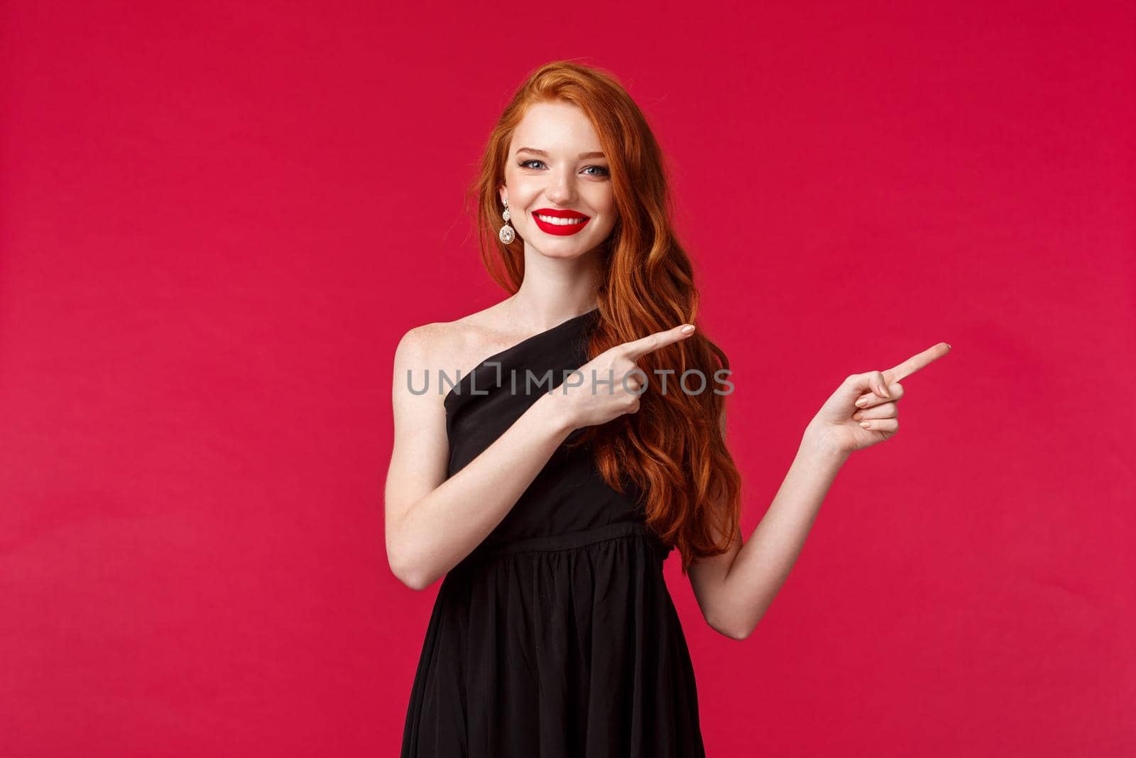Celebration, events, fashion concept. Portrait of sensual happy gorgeous redhead woman in black slim elegant dress, pointing fingers right and smiling, wear makeup, black background.