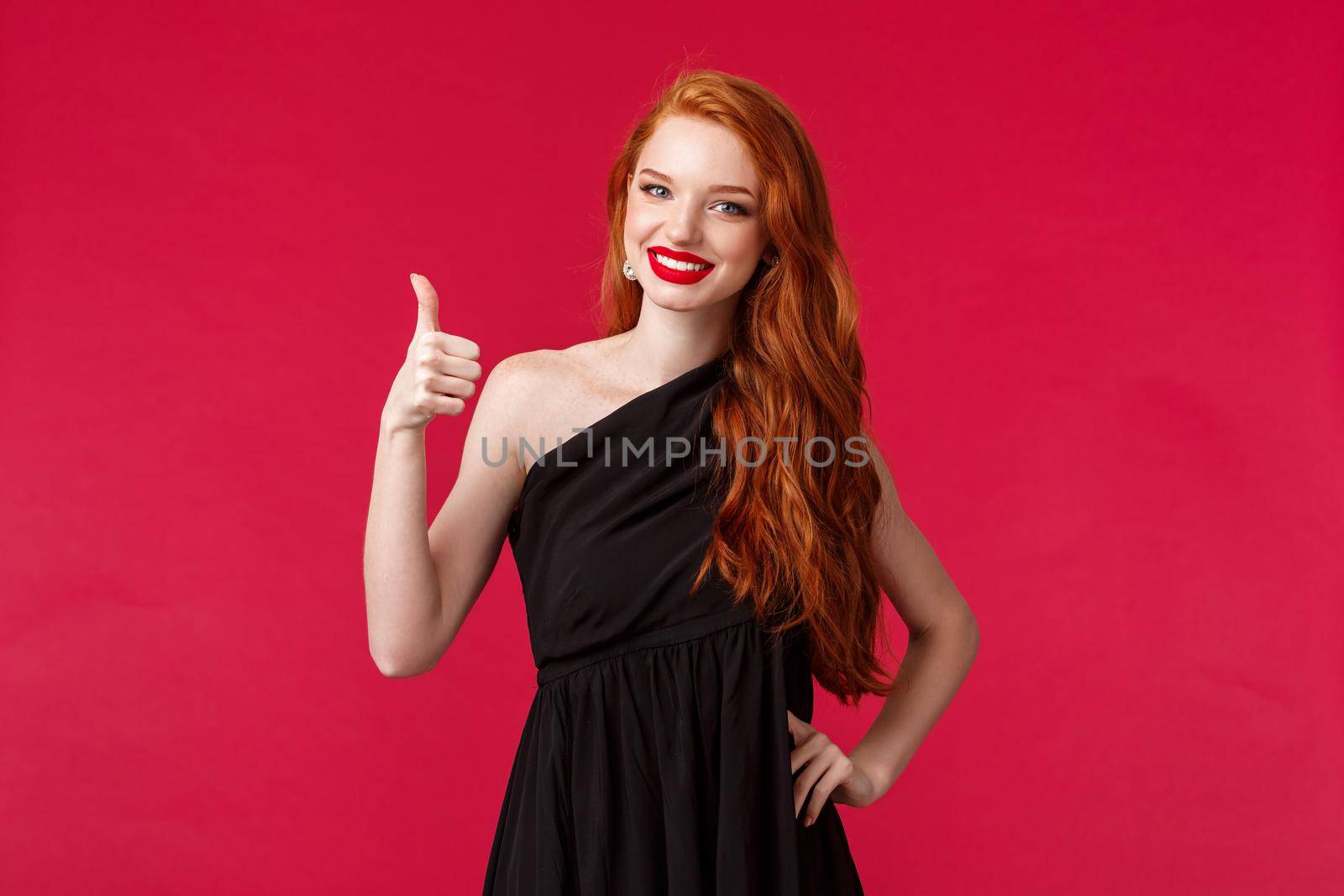 Fashion, luxury and beauty concept. Portrait of satisfied, assertive redhead elegant girl in black dress, show thumbs-up in approval or like gesture, smiling approvingly, red background by Benzoix