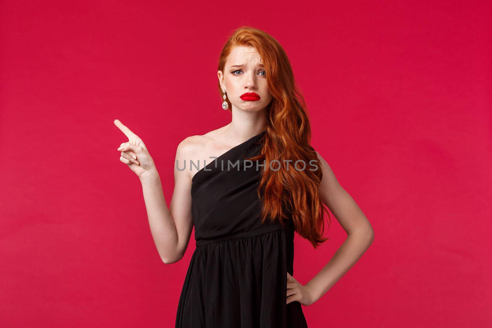 Beauty, fashion and women concept. Upset and displeased young gloomy redhead girl sighing and frowning unhappy pointing left look camera, express sadness wear black evening dress.