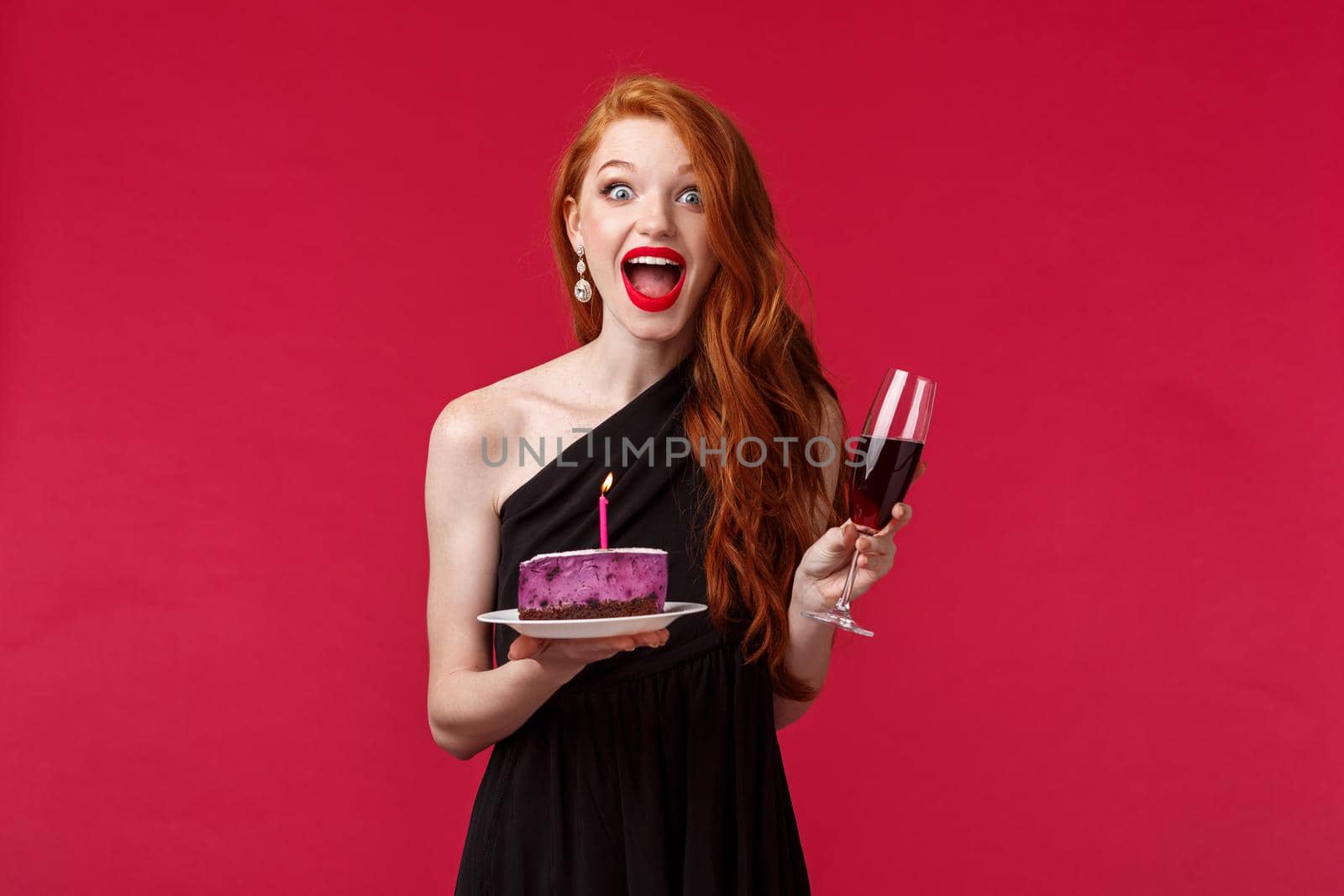 Portrait of excited and amused happy redhead b-day girl holding cake with lit candle and trying make wish blow it for dream come true, celebrating at party drinking wine, red background by Benzoix